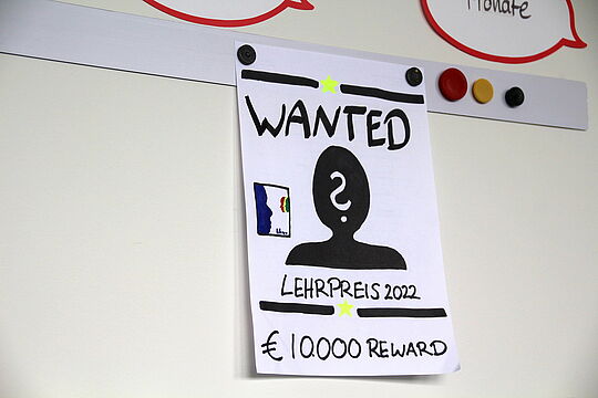 Illustration an einer Magnetwand. Plakat mit dem Titel "Wanted". Darunter eine anonyme, schwarze Gestalt, über deren Gesicht ein weißes Fragezeichen liegt. Neben ihr die Lehrpreis-Trophäe. Darunter die Aufschrift "Lehrpreis 2022, 10.000€ Reward".