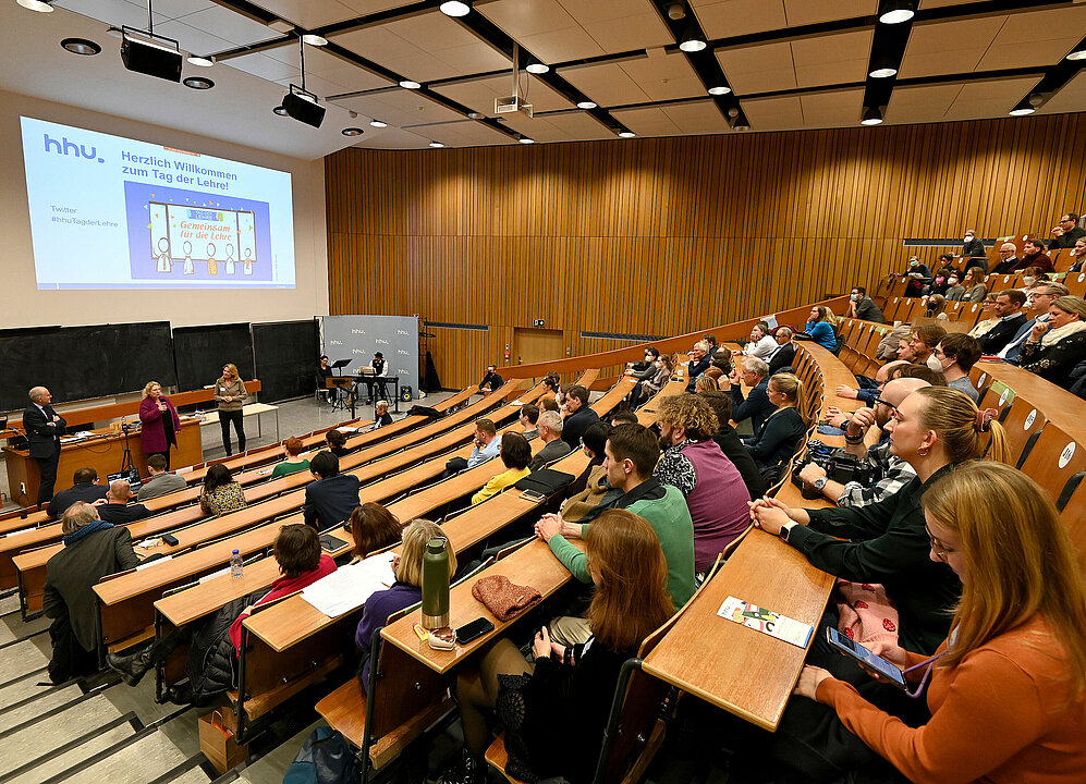 Blick in den gefüllten Hörsaal 2D an der HHU. Vor dem Rednerpult stehen Rektorin Prof. Dr. Anja Steinbeck, Prorektor Prof. Dr. Christoph Börner und Natalie Böddicker. Über ihnen eine Präsentation mit der Überschrift: Herzlich Willkommen zum Tag der Lehre.