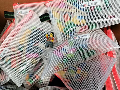 Foto von einer Lego-Frauenfigur mit schwarzen halblangen Haaren, die auf einem Haufen durchsichtiger Plastikbeutel mit orangenen Reissverschluss liegt. In den Plastikbeuteln sind lose Legosteine zu erkennen. Auf den Beuteln kleibt ein Aufkleber des Service-Center für gutes Lehren und Lernen (SeLL).