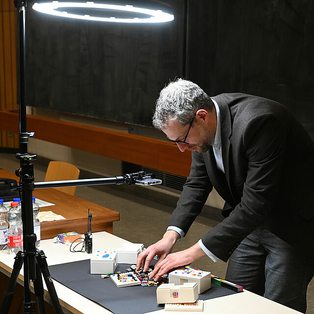 Foto von Peter Bernardi aus dem SeLL, der über einen Tisch gebeugt steht und Legosteine zusammenbaut. Über ihm auf den Tisch gerichtet ist eine Kamera mit Beleuchtung, über die die Szene auf der Leinwand übertragen wird.