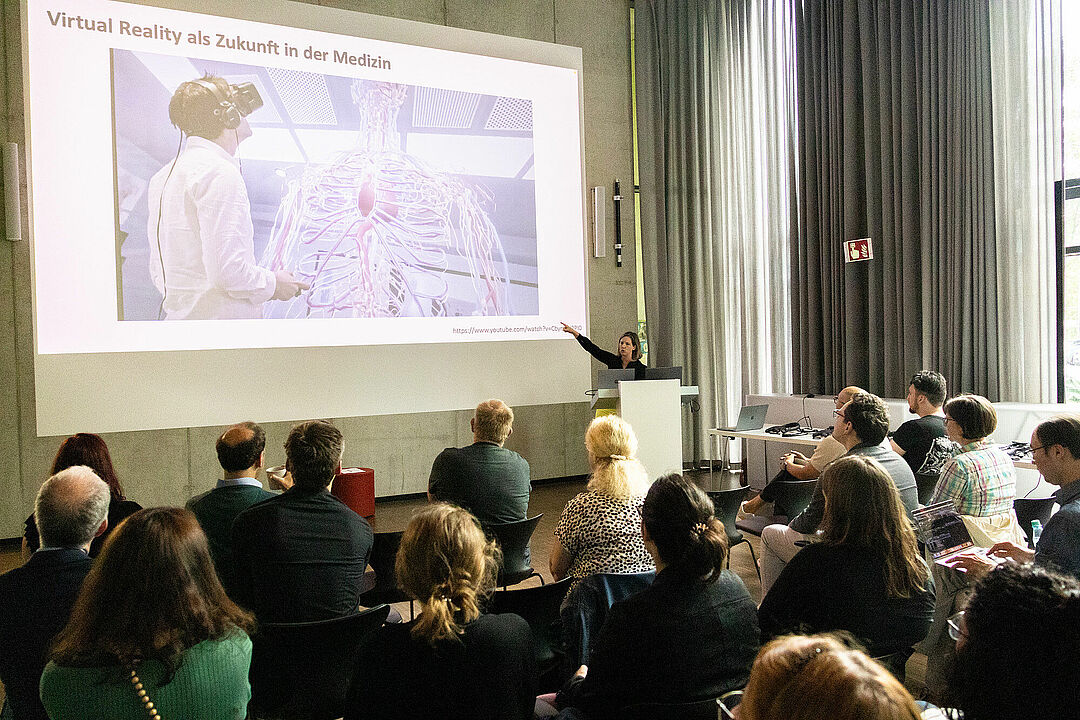 Ein Vortragssaal mit Publikum und einer Person am Stehpult, auf der Beamer-Leinwand ist eine Präsentation zu sehen. Auf der Folie steht: "Virtual Reality als Zukunft in der Medizin", abgebildet ist eine Person mit VR-Brille vor einem anatomischen 3D-Modell 