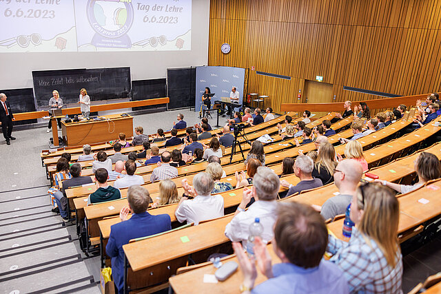 Besucher*innen des Tag der Lehre 2023 warten im Hörsaal 2A  gespannt auf ihren Plätzen auf den Beginn des Programms. Hinter einem Rednerpult stehen (von links nach rechts) Prof. Dr. Christoph J. Börner, Prorektor, Rektorin Anja Steinbeck und Natalie Böddicker.